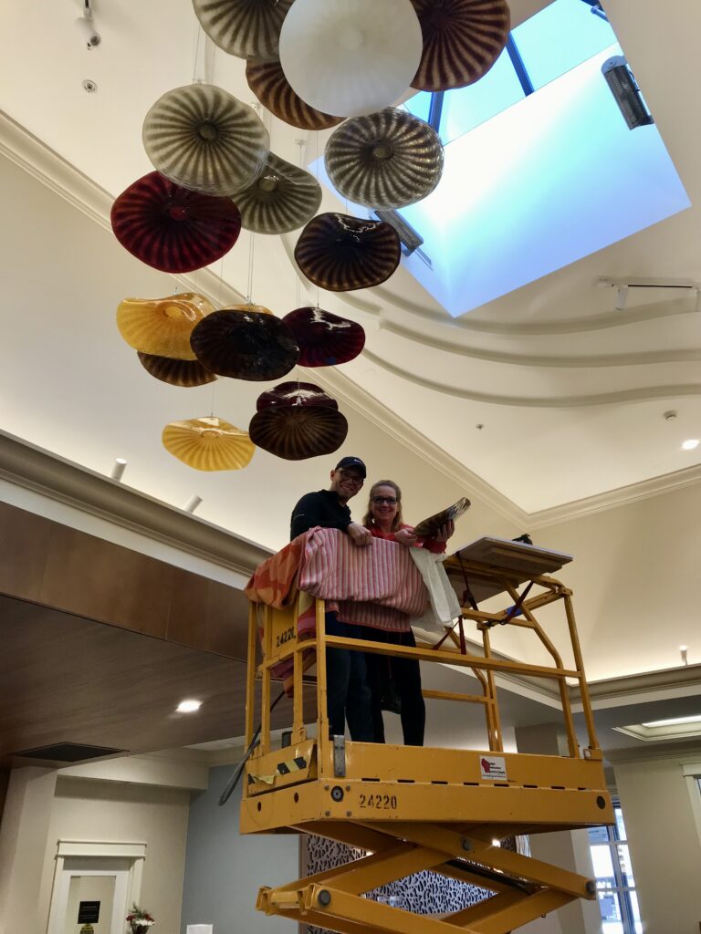Photo of Douglas and Renee Sigwarth standing on a lift underneath a hanging art installation.